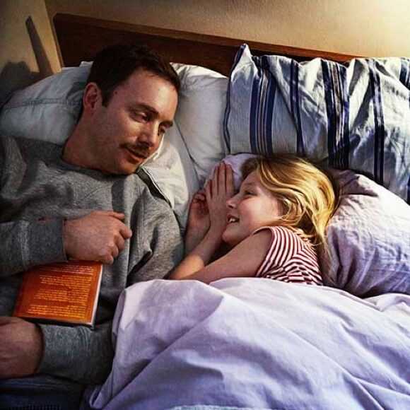 Fotografi: En pappa och en dotter ligger i sängen och pratar. I handen håller pappan en godnattsaga.