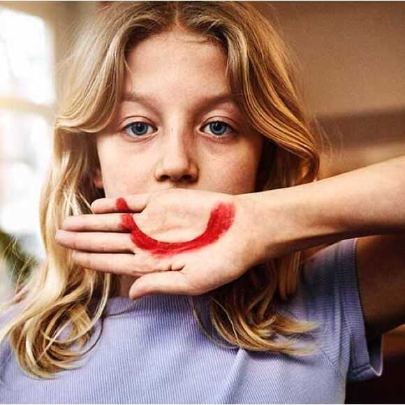 Fotografi: En sorgsen 10-årig flicka håller handen framför munnen. På handen har hon målat en stor röd glad mun.