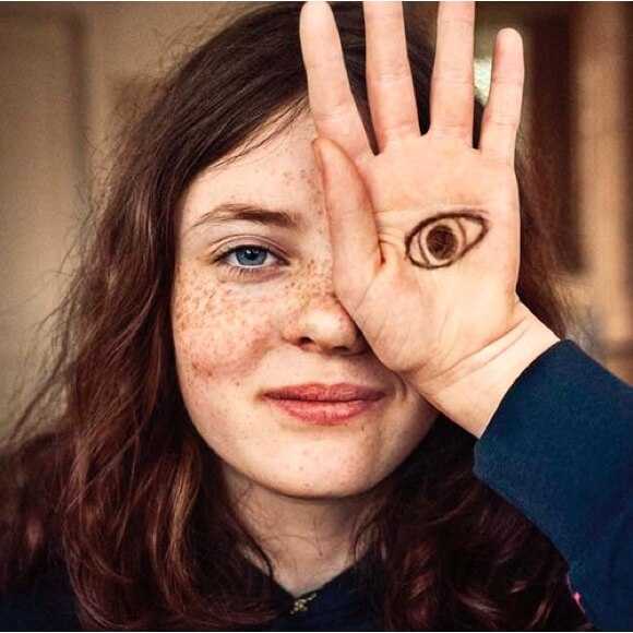 Fotografi: En flicka i tonåren ler mot kameran och håller ena handen framför ena ögat. I handen har hon ritat ett öga som tittar.