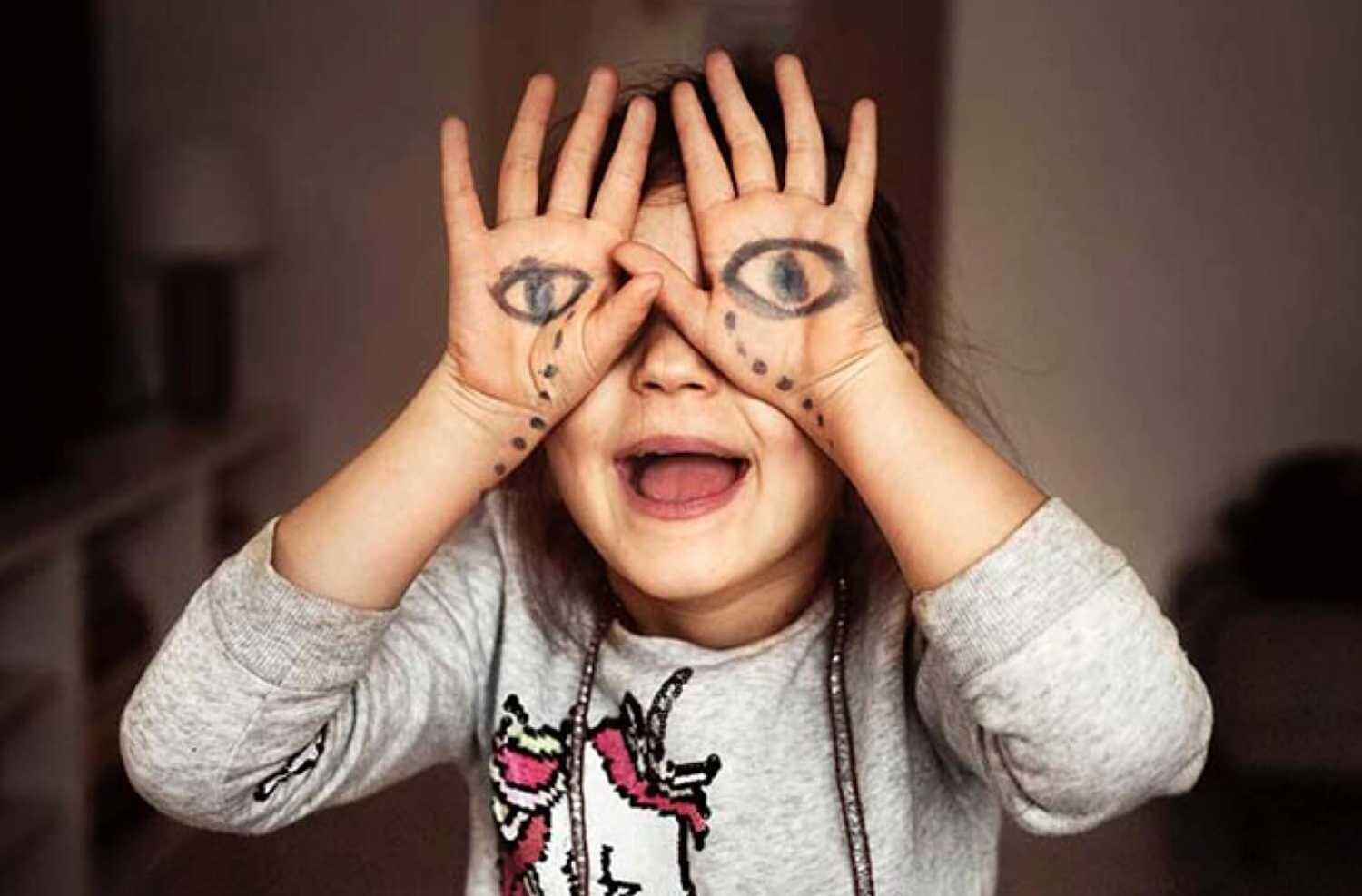 Fotografi: En glad 9-årig flicka som håller baksidan av händerna framför ögonen. På handflatorna har hon ritat ledsna ögon som gråter