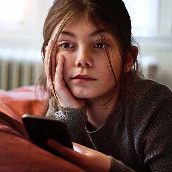 Fotografi: En fundersam 10-årig flicka ligger i soffan. I handen har hon en telefon, men hon tittar inte på den utan hennes blick tittar ut i det tomma intet.